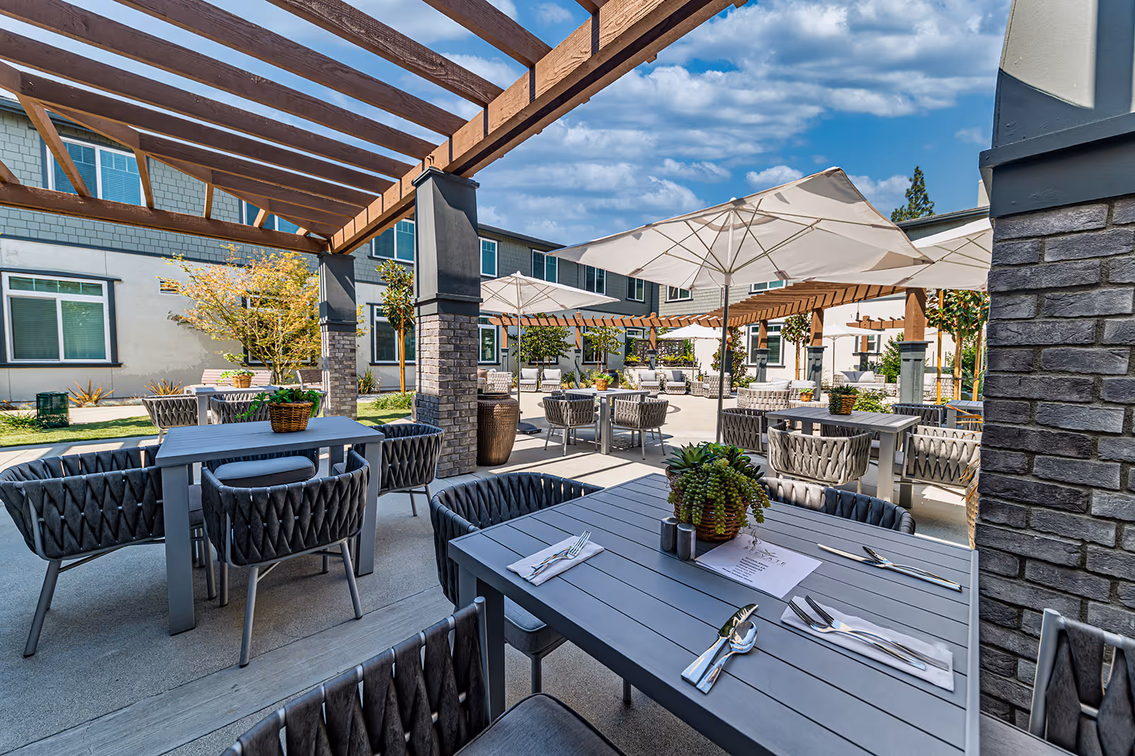 Outdoor patio area at Allara Senior Living with multiple tables and chairs arranged under large white umbrellas and wooden pergolas. The space is decorated with potted plants and surrounded by a building with windows. The sky is partly cloudy.