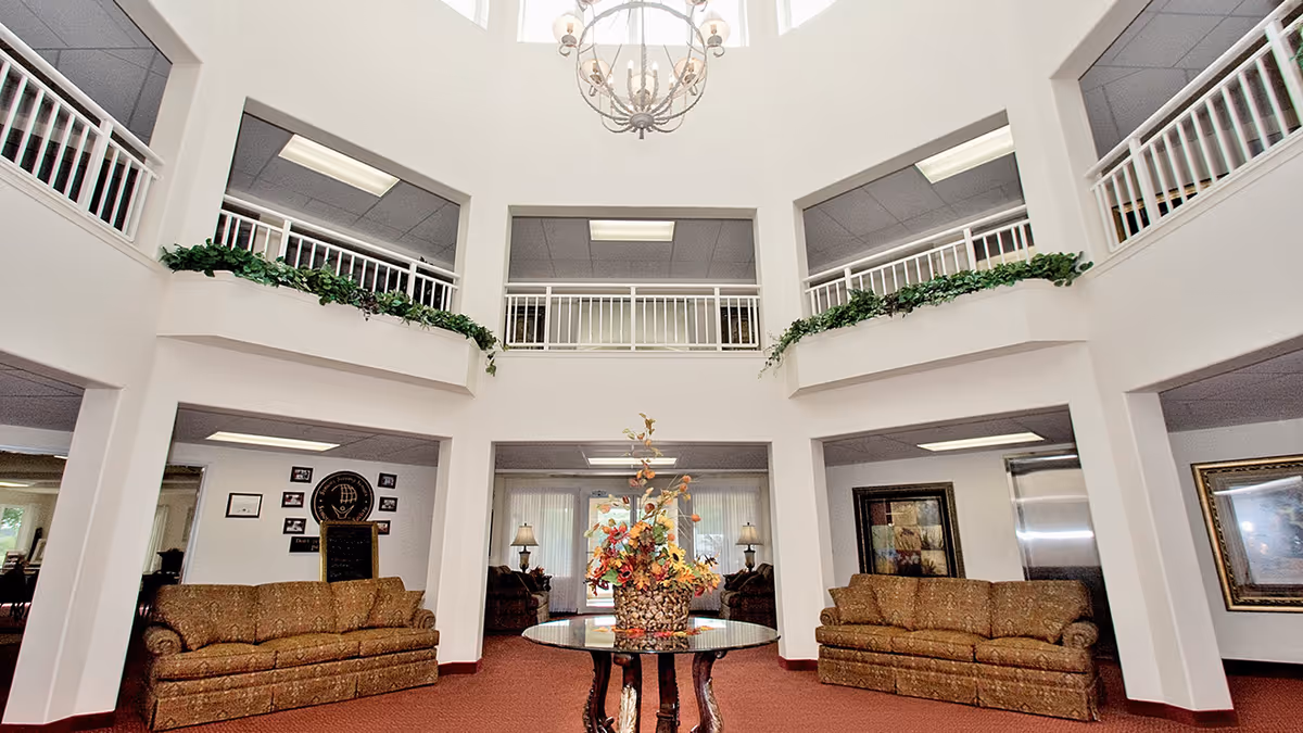 Spacious two-story atrium-style common lounge with sofas, a central table topped by a floral arrangement, upper balconies, and a chandelier.