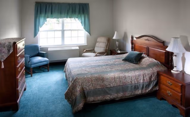 Sunlit bedroom with a wooden bed, matching nightstands and dresser, teal carpet and a window with a valance