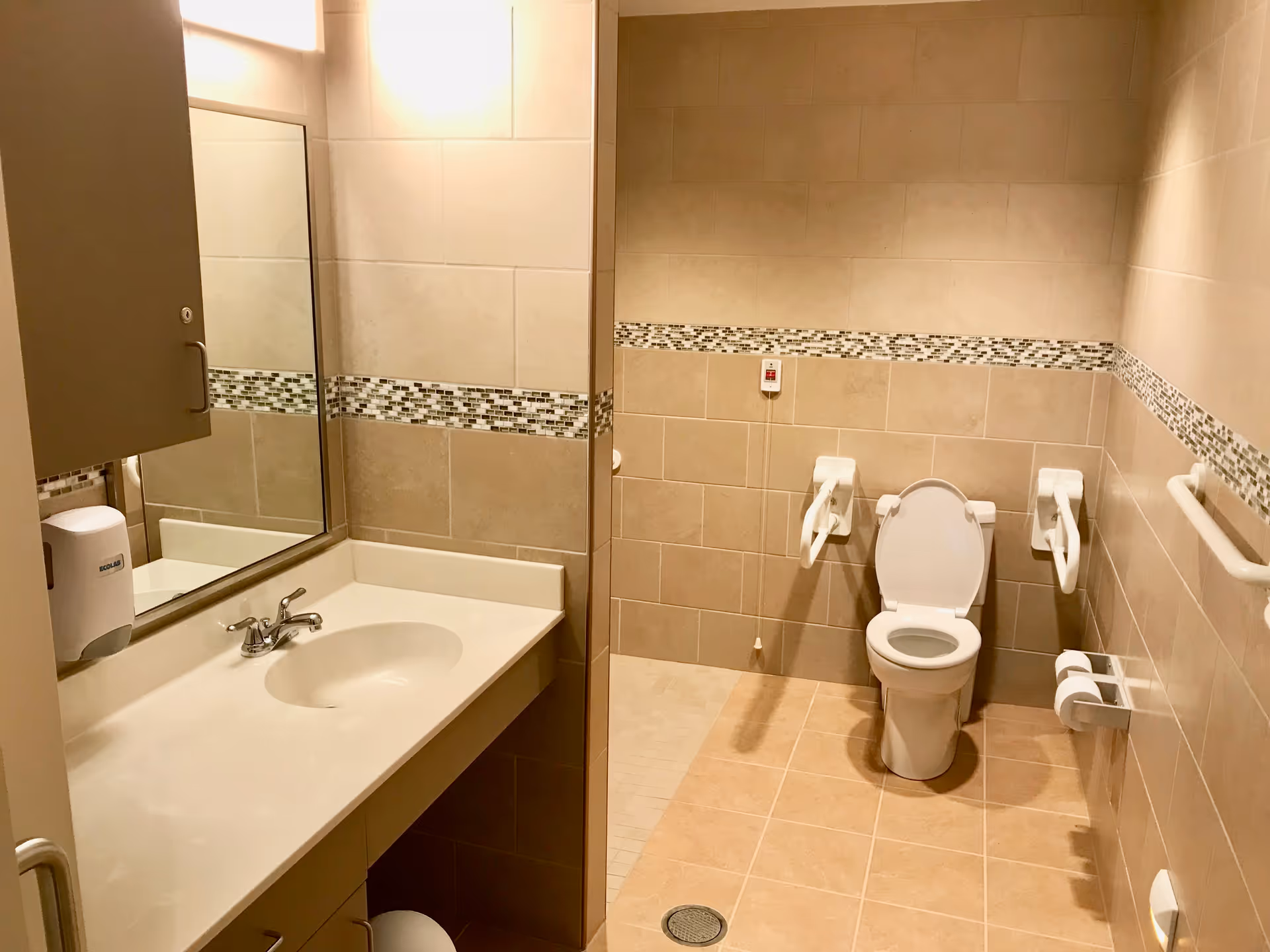 A clean, accessible bathroom with beige tiled walls and floor. The bathroom features a white sink with a countertop and a mirror above it on the left side. On the right side, there is a toilet with support rails on both sides and a pull cord emergency alarm on the wall behind it. A soap dispenser is mounted on the wall near the sink.