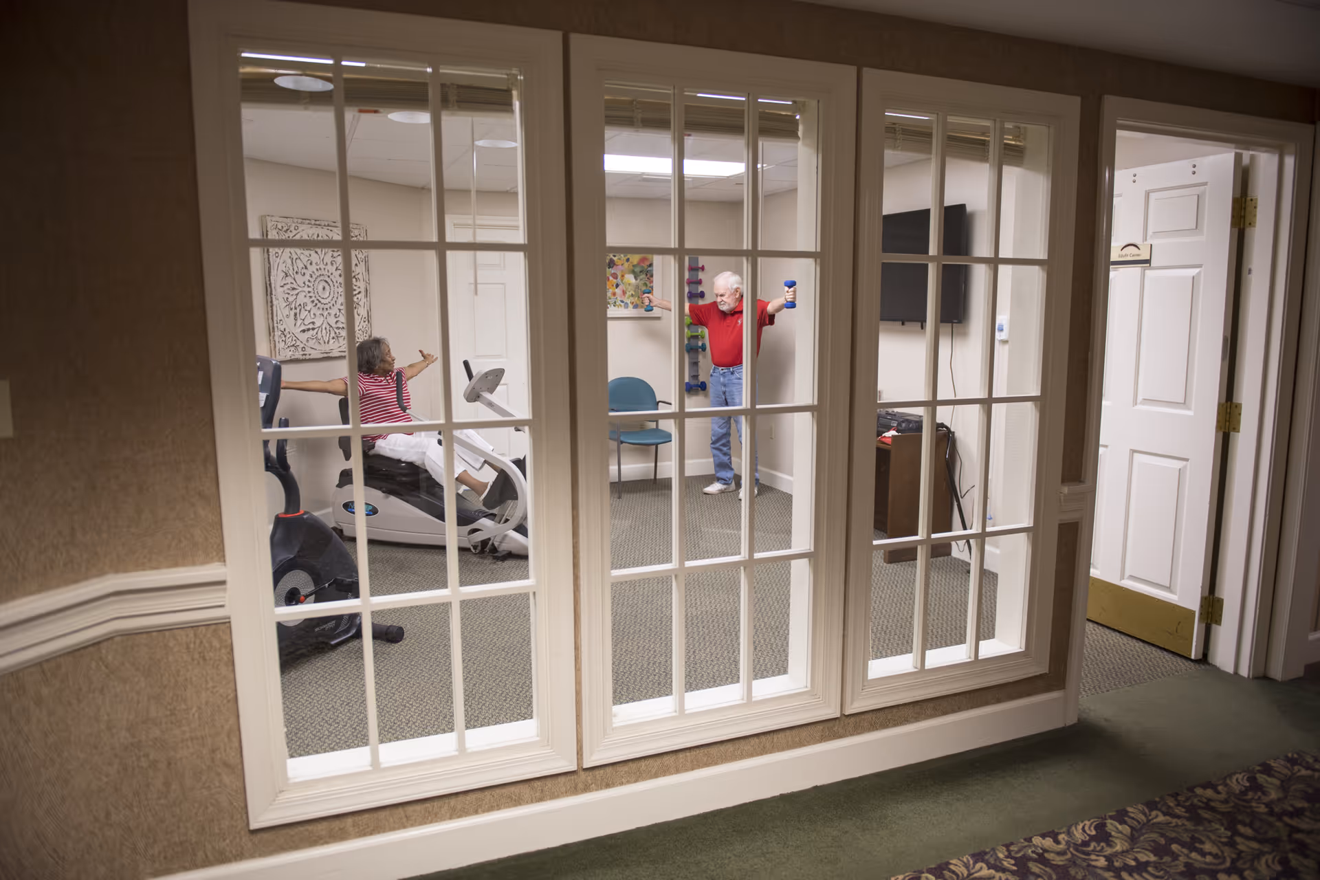 View through large glass windows into a fitness room where an elderly woman is using a recumbent exercise bike and an elderly man is lifting small dumbbells. The room has beige walls, carpeted floor, and some wall decorations. The door to the room is open and labeled 'Exhibit Center.'