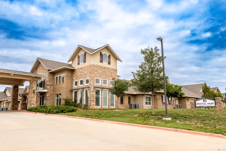 Exterior view of Bristol Park at Amarillo Assisted & Living Memory Care facility showing a large building with stone and beige siding, multiple windows, a covered entrance, a driveway, green lawn, trees, and a sign with the facility name.