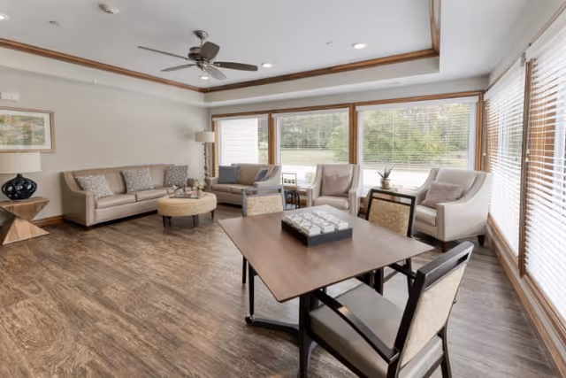 A bright and spacious living room with large windows allowing natural light to fill the space. The room features a ceiling fan, a beige sofa with patterned cushions, two armchairs, a round ottoman coffee table, and a wooden table with four chairs. The floor has a wood-like finish, and there are decorative elements including a lamp, a framed picture, and small plants on side tables.