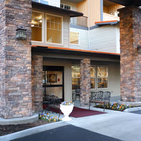 Entrance of a senior living facility with stone pillars, a red carpet leading to glass doors, outdoor benches, and flower beds on either side.