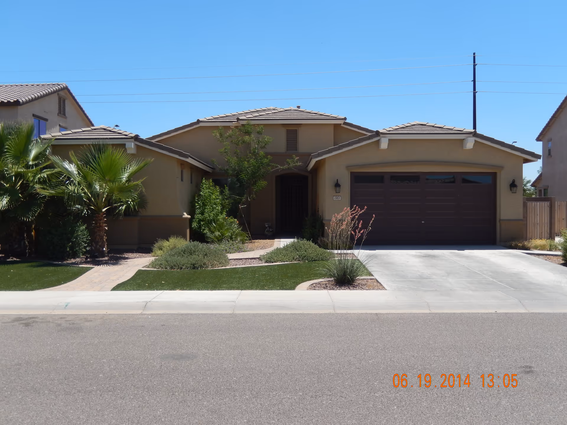 Front exterior view of a single-story house with a tiled roof, a two-car garage with a brown door, a paved driveway, and a landscaped front yard featuring palm trees, bushes, and a small tree. The sky is clear and blue.