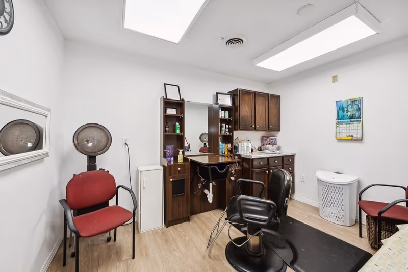 Interior of a small salon or beauty room with a hair washing station, a black salon chair, two red chairs, wooden cabinets with various hair products, a calendar on the wall, and a laundry basket. The room has white walls and a light wood floor with bright overhead lighting.