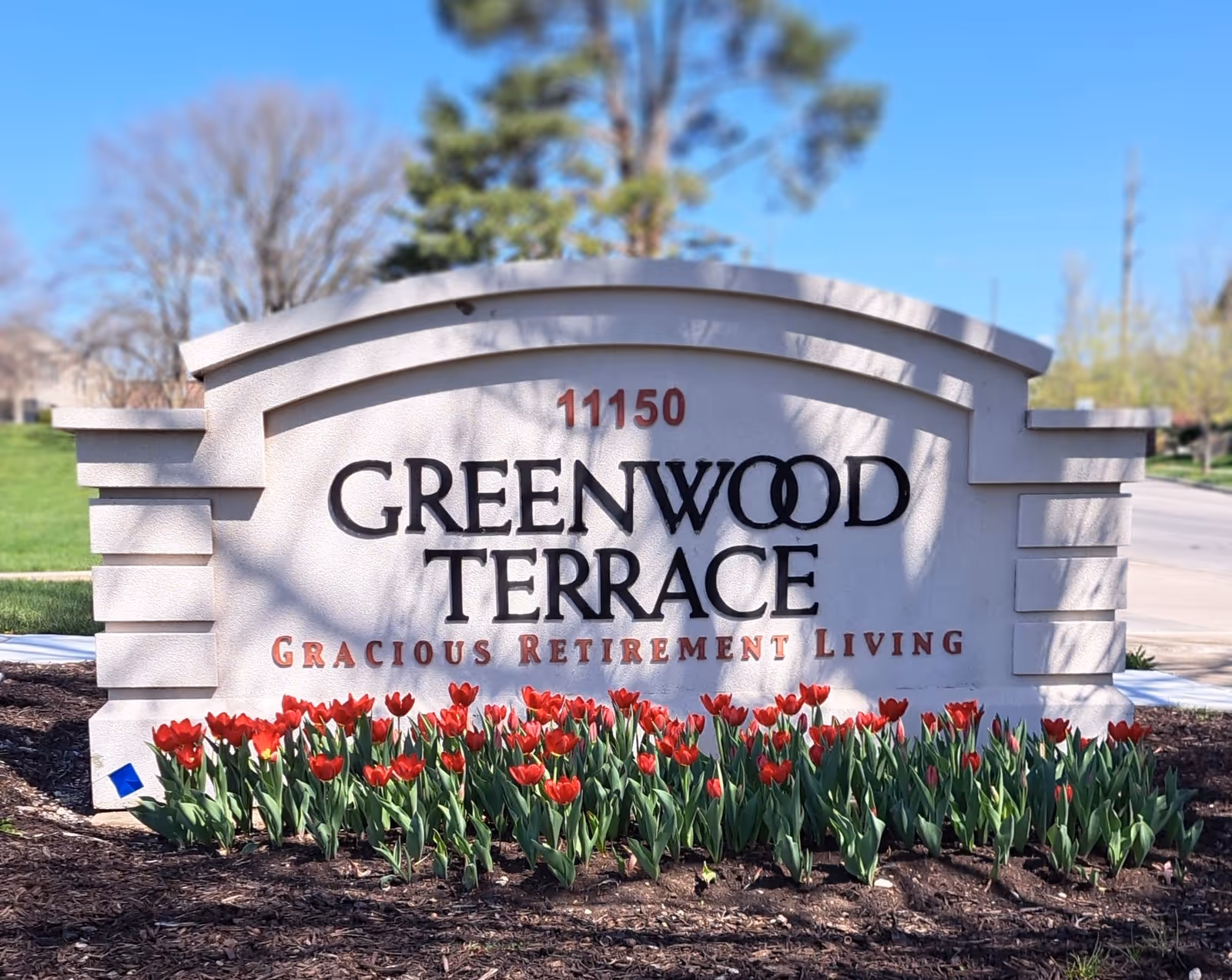 Outdoor stone sign for Greenwood Terrace retirement living with red tulips planted in front and trees and grass in the background under a clear blue sky.