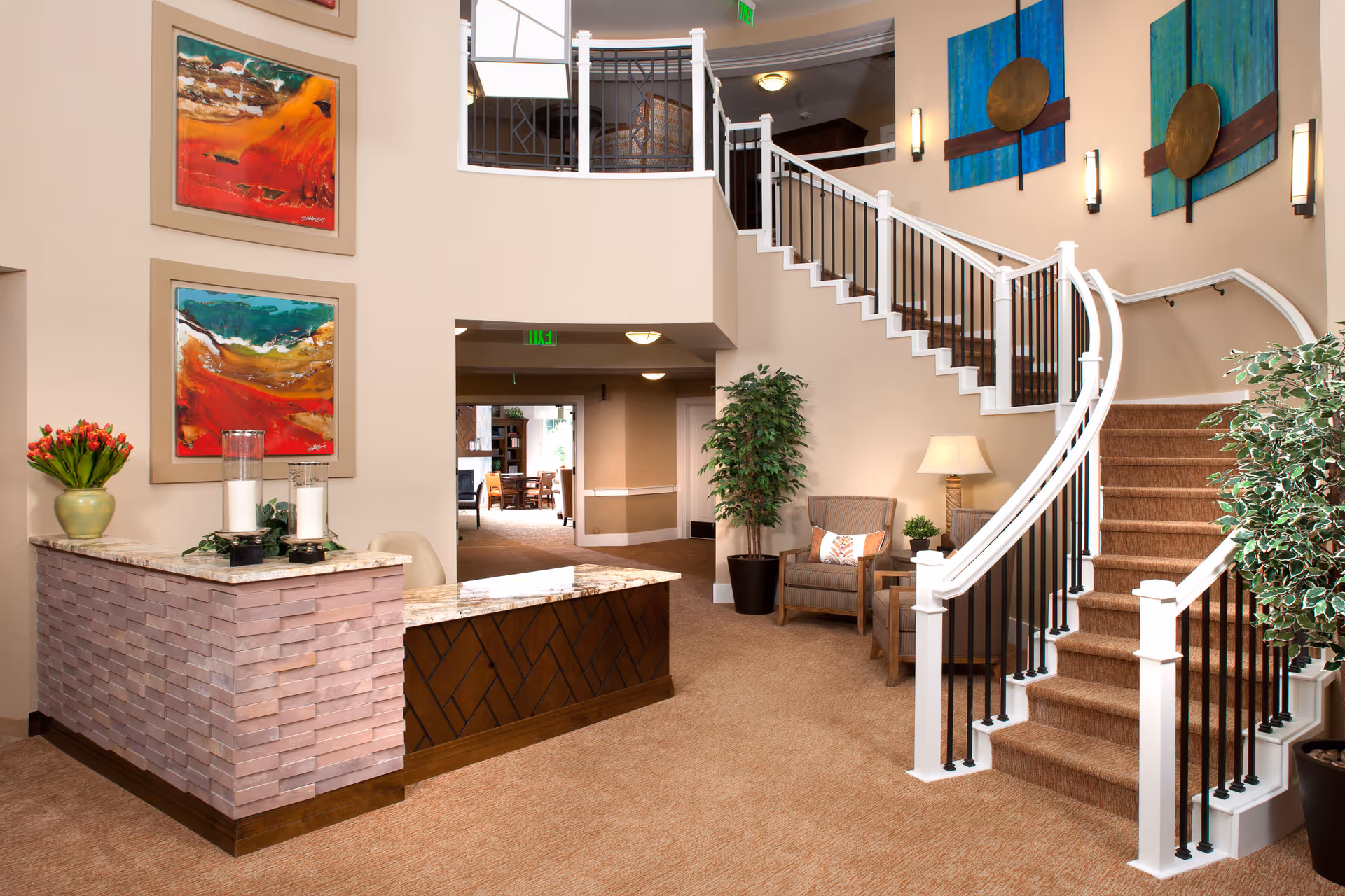 Interior view of a senior living facility lobby with a curved staircase featuring white railings and black balusters. The space includes a reception desk with a stone facade and marble countertop, decorated with a vase of red tulips and two large candles. There are colorful abstract paintings on the walls, a comfortable armchair with a pillow, a small side table with a lamp, and several potted plants. The area is well-lit with wall sconces and ceiling lights, and an open doorway leads to another room with tables and chairs.