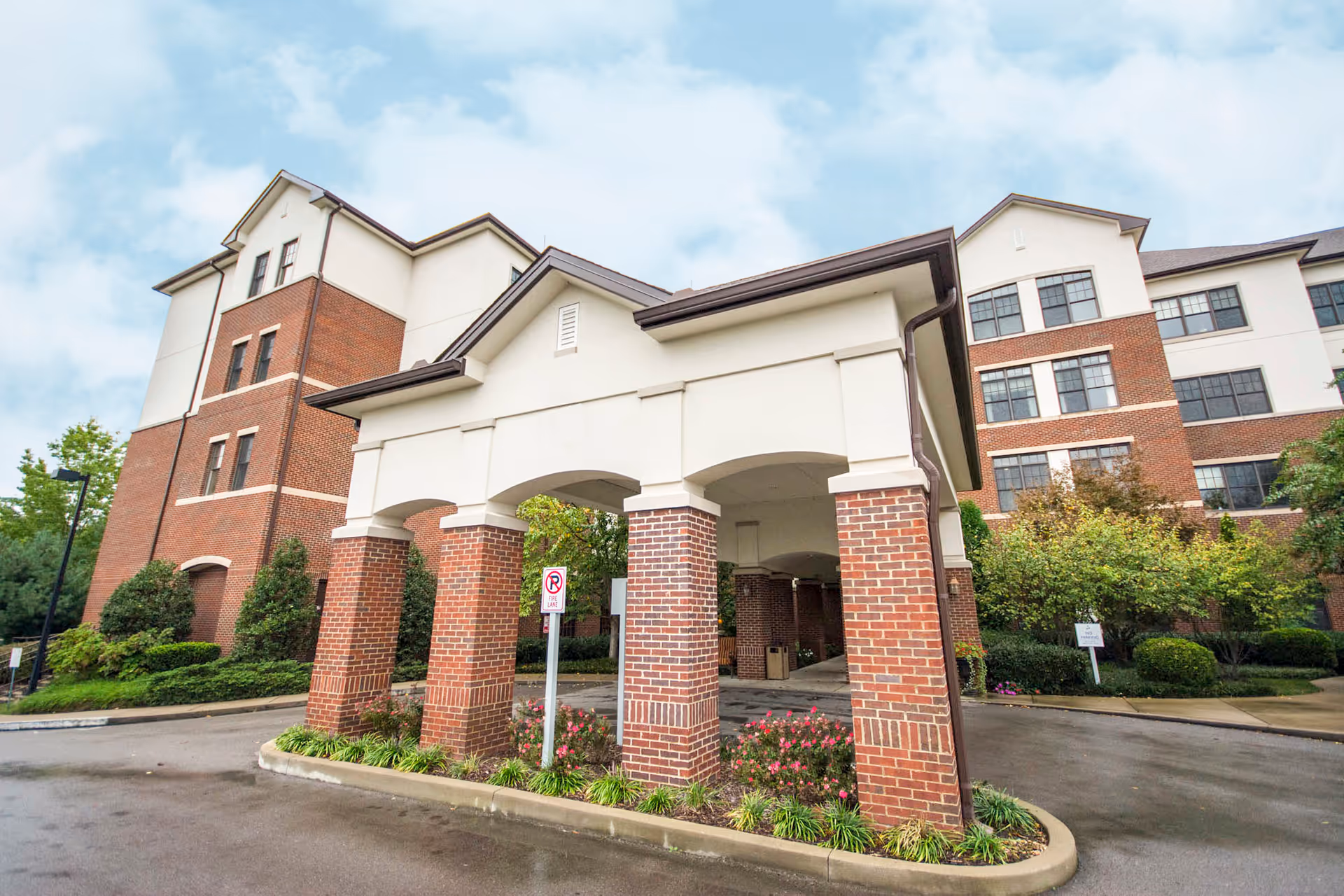 Exterior view of Brookdale Green Hills Cumberland senior living facility showing a covered entrance with brick pillars and a multi-story building with brick and white facade under a partly cloudy sky.