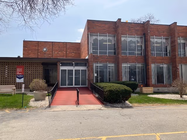 Front exterior of a two-story brick building with a red wheelchair ramp leading to glass entrance flanked by large windows and shrubs.