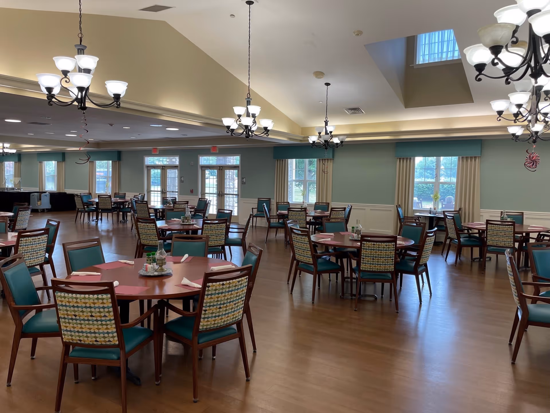 A spacious dining room with multiple round tables and chairs arranged neatly. The chairs have patterned backs and teal seats. The room features large windows with curtains, wooden flooring, and several chandeliers hanging from a high ceiling with recessed lighting. There are doors leading outside and some decorative items on the tables.