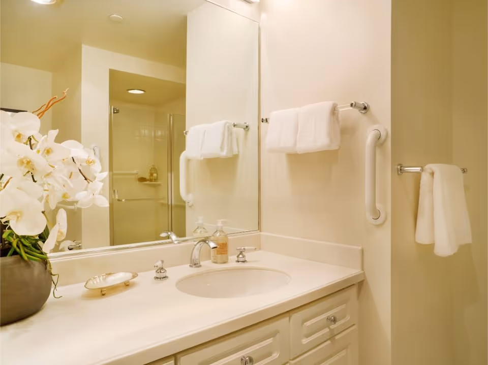 A clean bathroom vanity with a white countertop, sink, and silver faucet. There are two soap dispensers on the counter and a decorative vase with white orchids. A large mirror is mounted above the sink, reflecting a glass-enclosed shower with white tiles. White towels hang on towel bars and grab bars are installed on the walls for support.