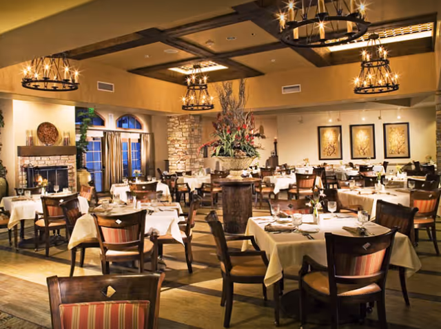 A warmly lit dining room with multiple tables covered in white tablecloths, set with plates, glasses, and napkins. The room features wooden chairs with striped cushions, a stone fireplace, large windows with curtains, decorative wall art, and chandeliers hanging from a ceiling with wooden beams. A large floral arrangement is placed on a central pedestal.