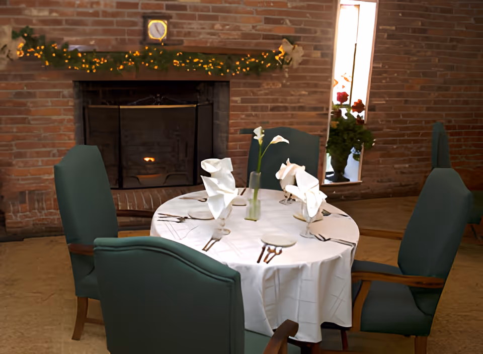 A round dining table set for four with white tablecloth and folded white napkins in glasses, placed in front of a brick fireplace decorated with a garland of greenery and lights. Four green upholstered chairs surround the table, and a tall narrow window with flowers is visible on the right side.