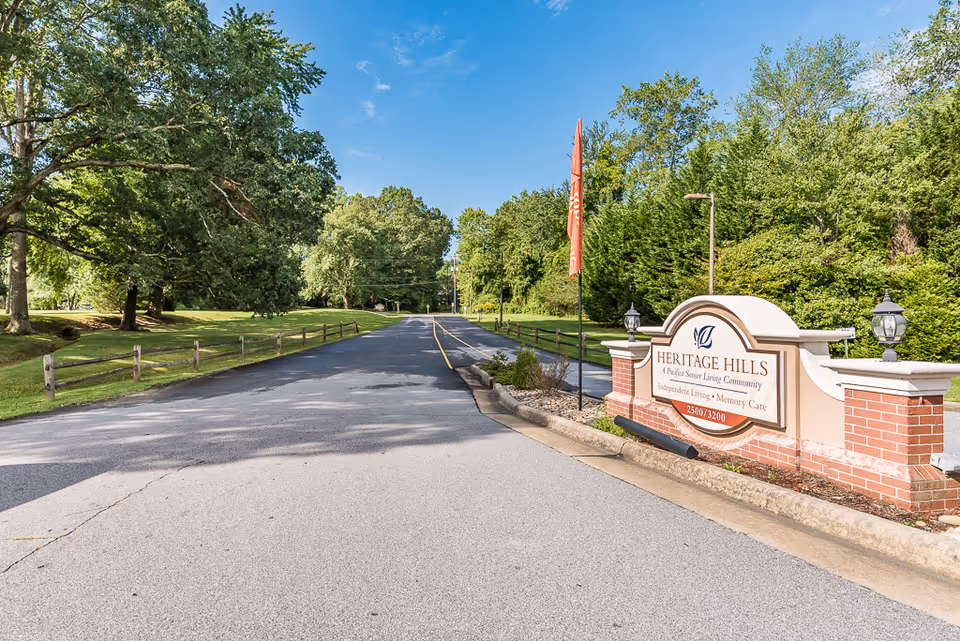 Entrance driveway and landscaped sign reading 'Heritage Hills' for a senior living community surrounded by trees under a blue sky.