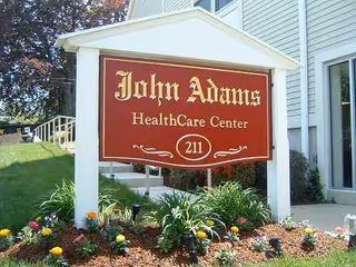 A sign reading John Adams Healthcare Center stands outside a light-colored building entrance.