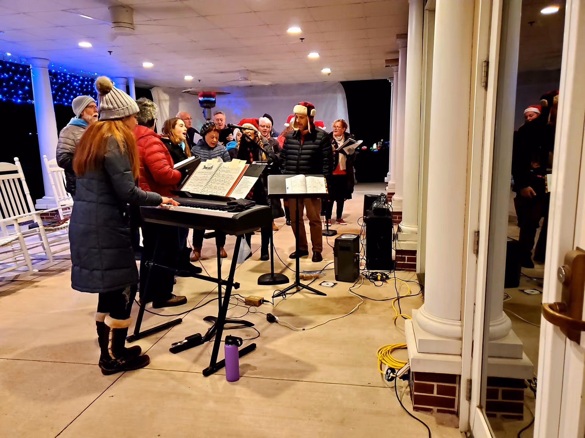 A group of people dressed in winter clothing, some wearing Santa hats, are gathered on a covered outdoor patio at night. One person is playing an electronic keyboard while others are singing from songbooks. The area is decorated with blue string lights and white rocking chairs are visible in the background. There are columns supporting the patio roof and some audio equipment on the ground.