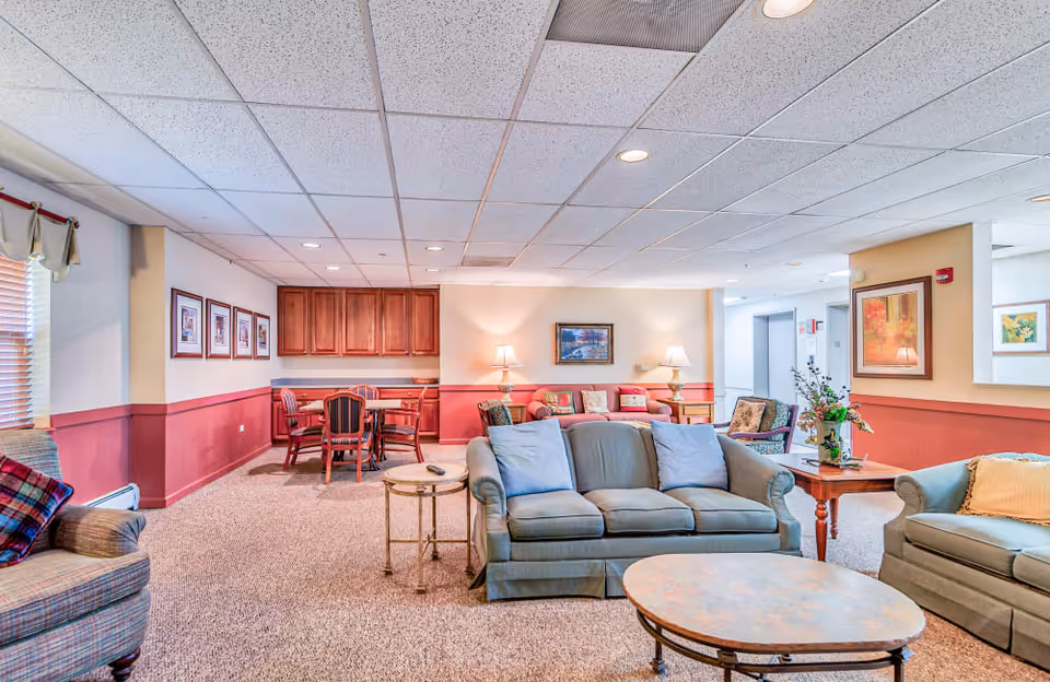 A spacious senior living facility lounge with sofas, coffee tables, and a dining area in the background.