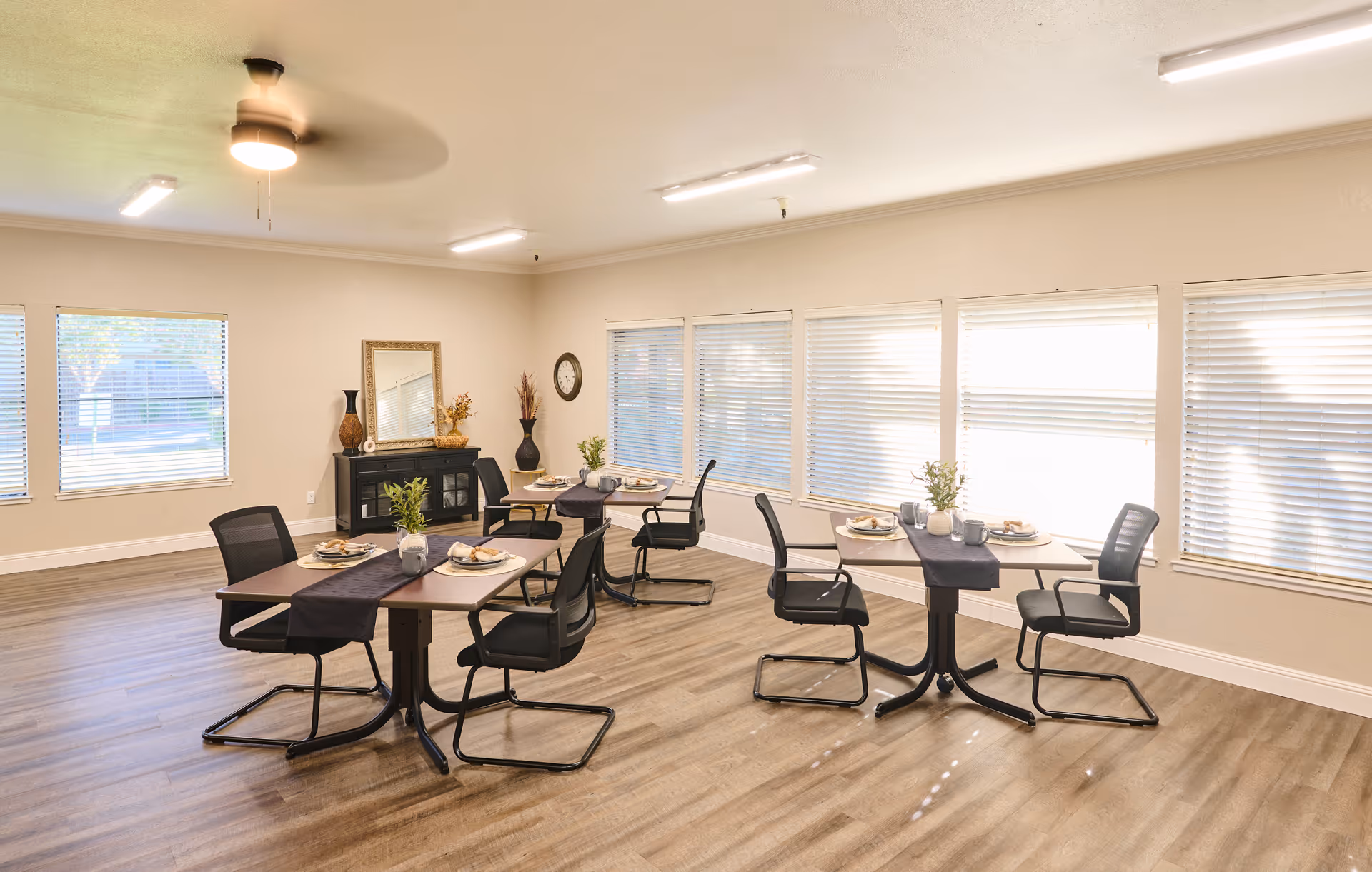 Communal dining room with several small tables set for meals, black chairs, large windows with blinds, and decorative accents.
