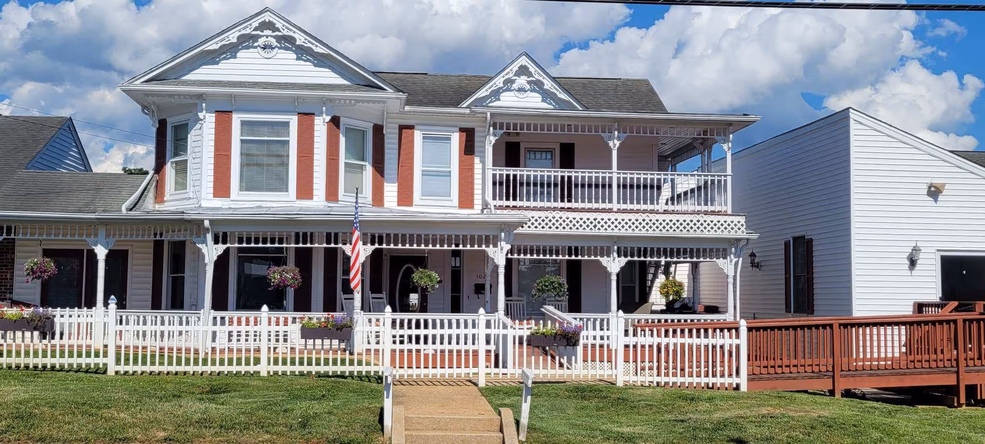 A large two-story white house with red shutters and decorative trim, featuring a wraparound porch with hanging flower baskets and an American flag in front. The house is surrounded by a white picket fence and has a wooden ramp on the right side. The sky is blue with scattered clouds.