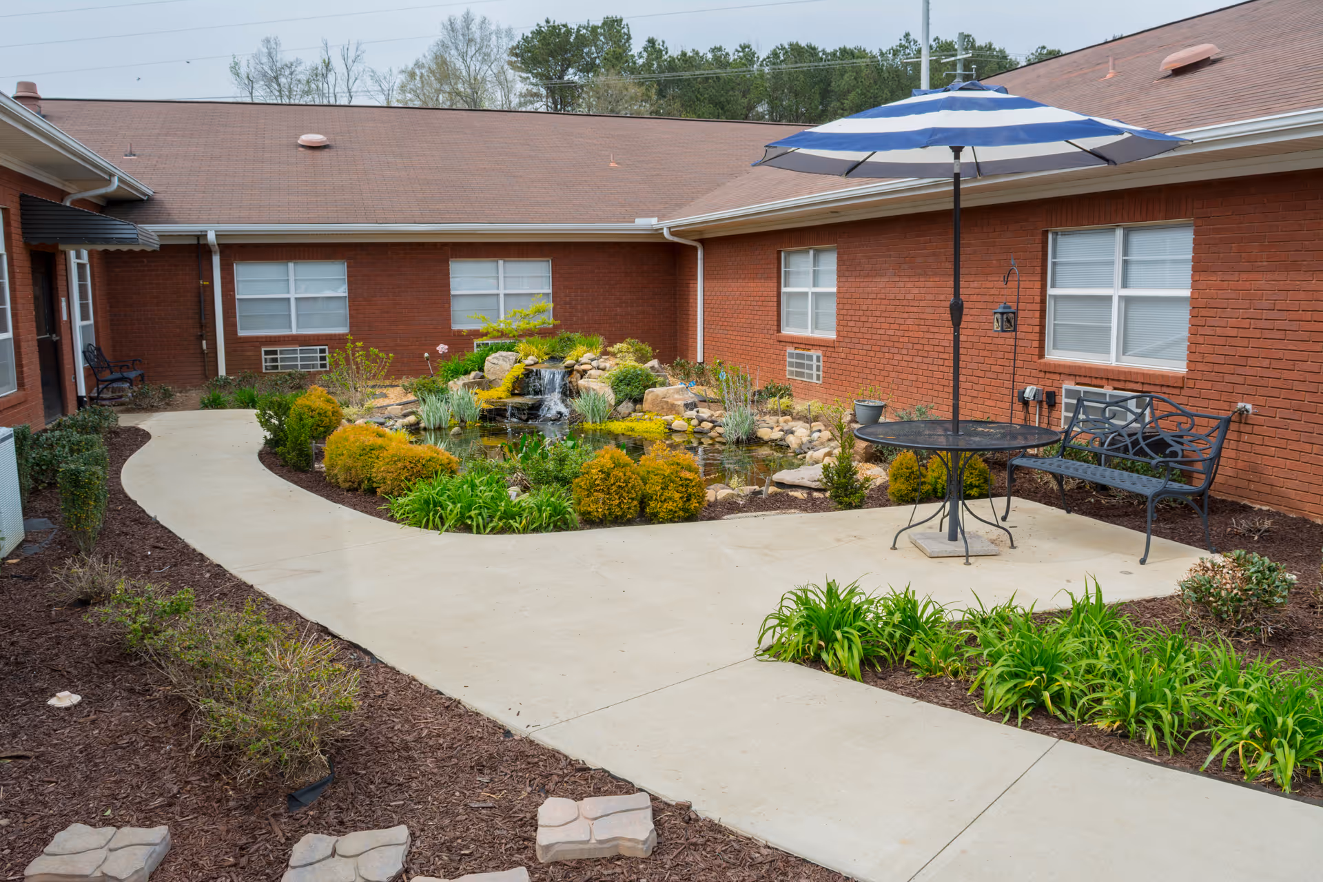 Outdoor courtyard area at Cleburne County Nursing Home featuring a small pond with a waterfall surrounded by greenery and shrubs. There is a concrete pathway curving around the pond, a metal bench, and a round table with a blue and white striped umbrella. The courtyard is enclosed by a single-story red brick building with several windows.