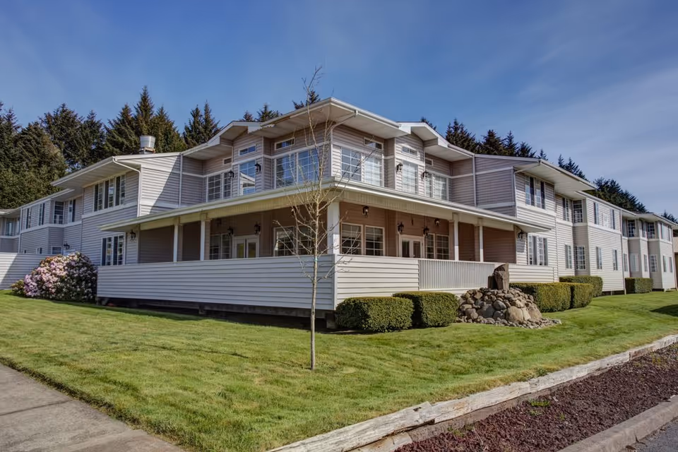 Two-story light-colored assisted living building with a covered wraparound porch, trimmed hedges and a green lawn under a blue sky.
