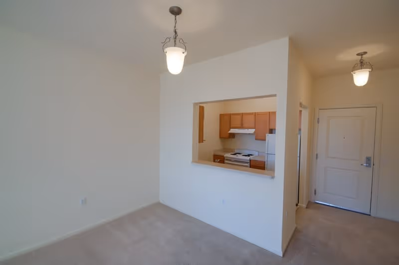 Empty interior room with beige carpet and white walls, featuring a pass-through window opening to a kitchen with wooden cabinets, white stove, and refrigerator. Two ceiling light fixtures are visible, and a white entrance door is seen in the background.