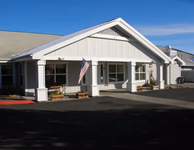 Single-story white senior living facility with a peaked roof, front entrance, and flag.