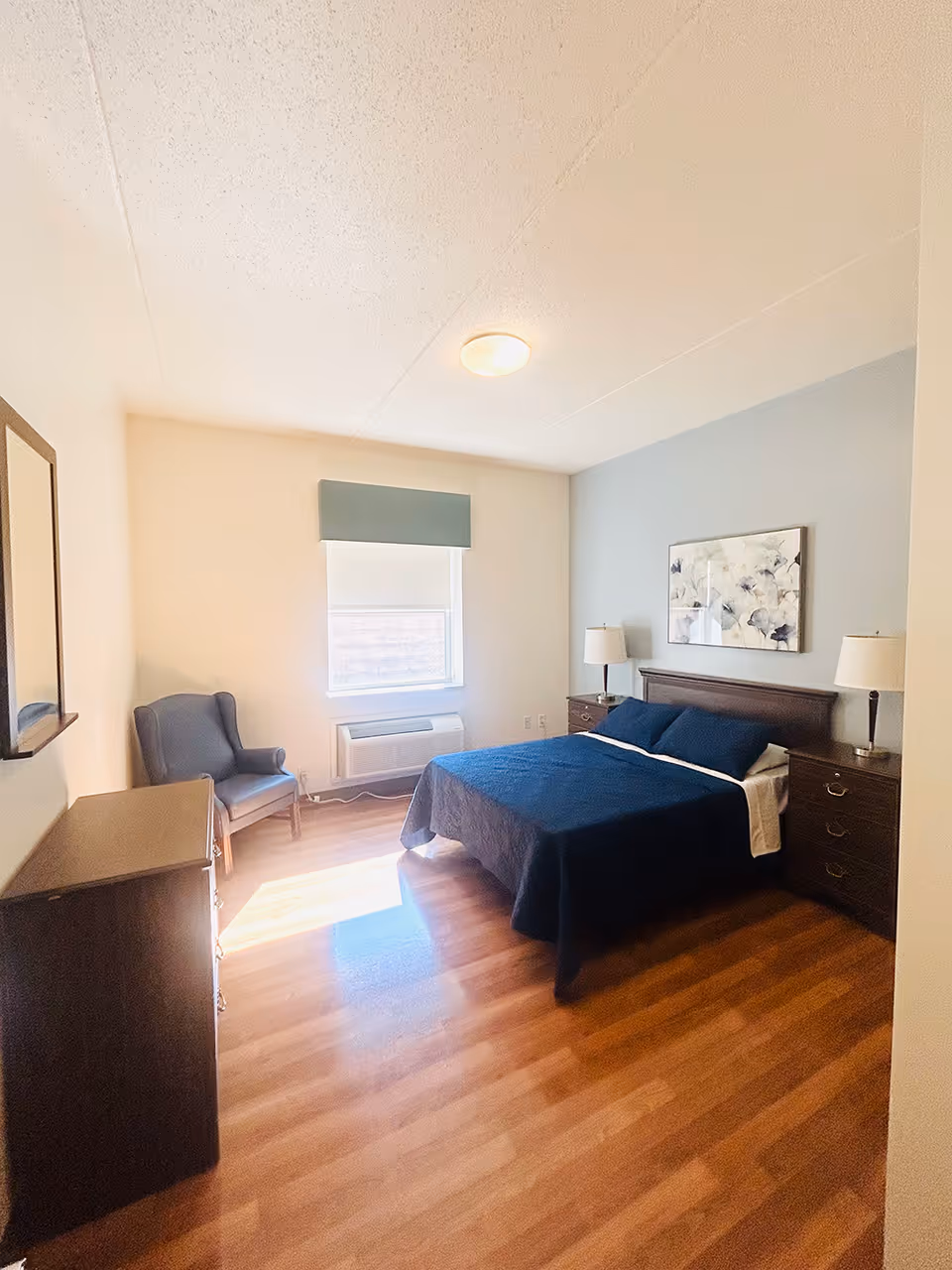 A bright bedroom with a wooden floor, a bed with dark blue bedding, two bedside tables with lamps, a framed floral artwork above the bed, a window with a blue valance, a blue armchair, and a wooden dresser with a mirror on the left wall.