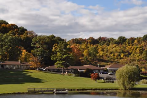 Autumn-colored trees frame a lakeside assisted living campus with low buildings and a green lawn.