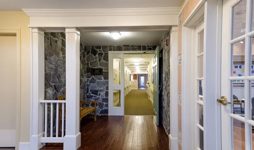 Interior corridor of a senior living facility with white columns, stone-accent walls, a wooden bench and doors opening to rooms.