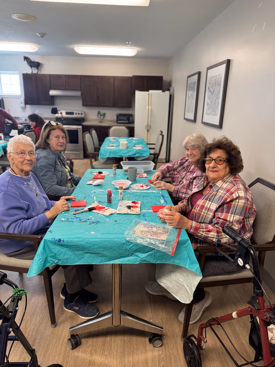 Four elderly women sitting around a table covered with a turquoise tablecloth, engaging in a craft activity with paint and brushes in a kitchen area. The room has wooden cabinets, a stove, a refrigerator, and framed artwork on the wall. Two walkers are visible near the women.