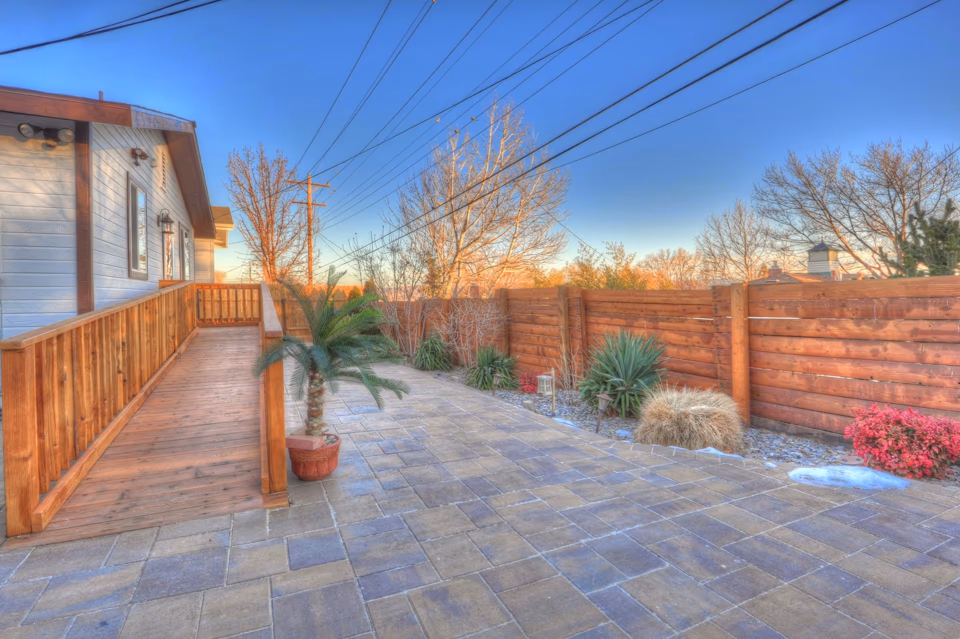 Outdoor patio area with a wooden ramp leading to a building on the left. The patio is paved with stone tiles and bordered by a wooden fence. Various plants and small bushes are planted along the fence. The sky is clear with visible power lines overhead.