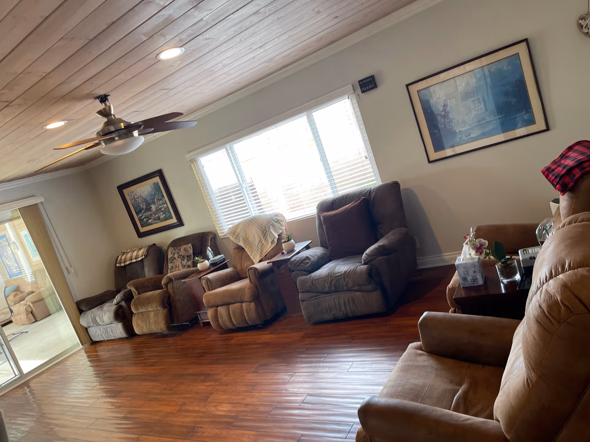 Sunlit living room with multiple recliner chairs along a wall, a ceiling fan, framed artwork, and hardwood floors.