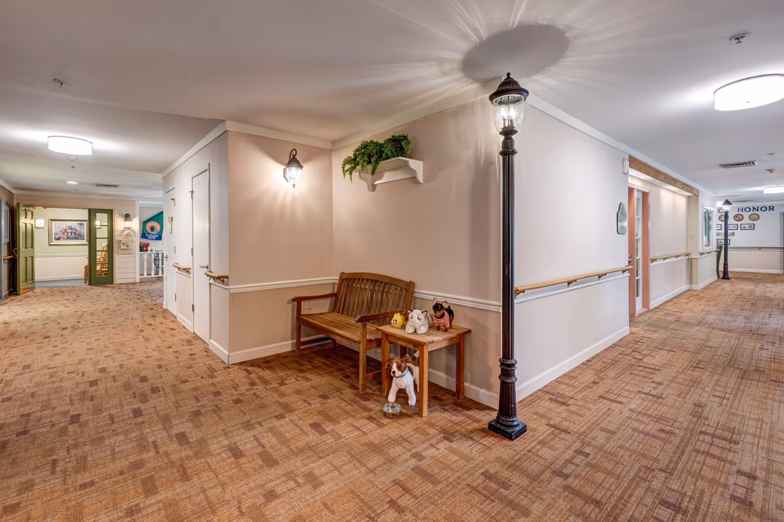 Interior hallway of Arden Courts - ProMedica Memory Care Community with beige walls, patterned carpet, wooden bench, small table with decorative items, wall-mounted lights, and a streetlamp-style floor lamp. The hallway has handrails along the walls and doors leading to rooms.
