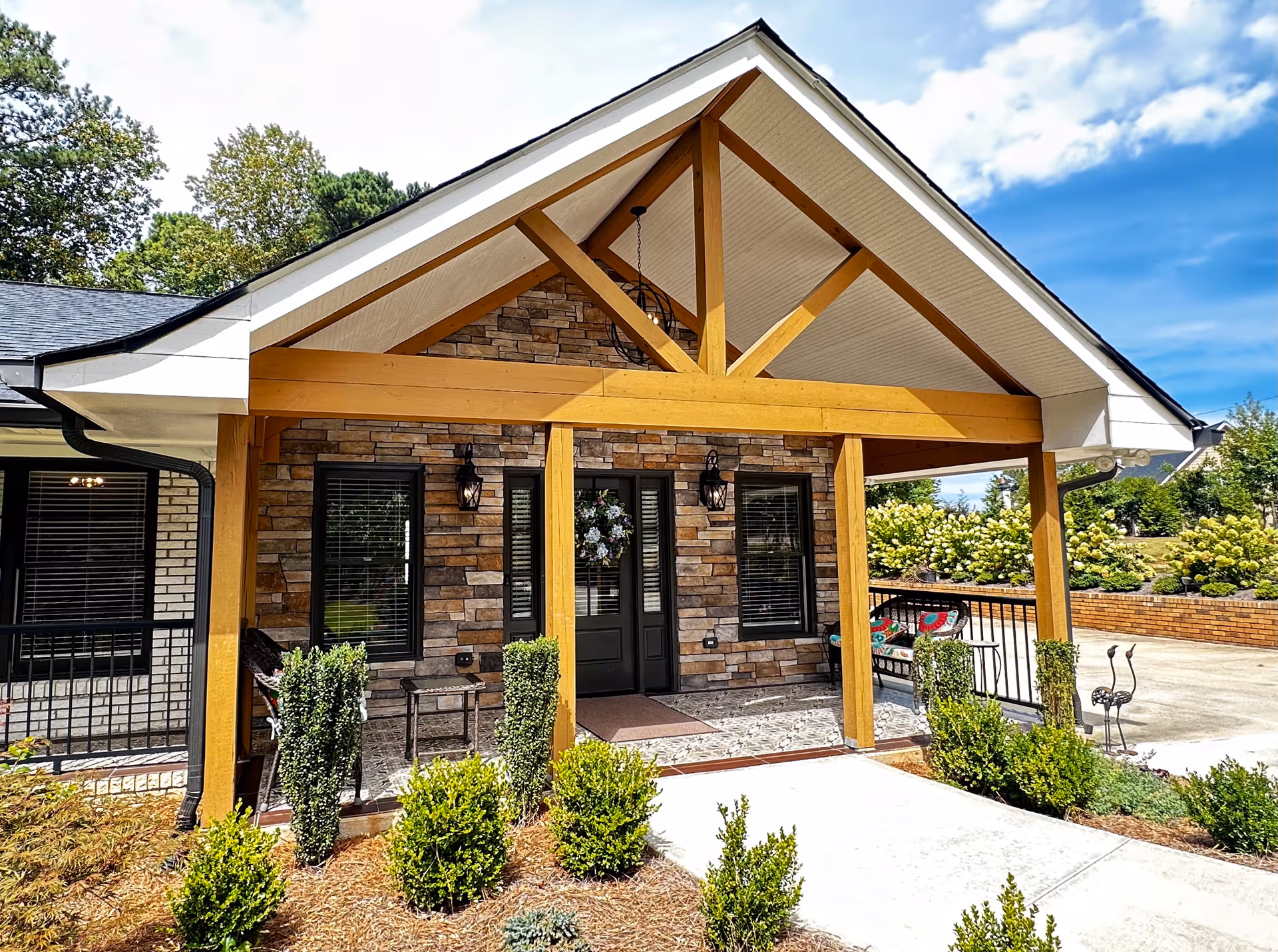 Front entrance of a senior living facility with a covered porch supported by wooden beams, stone facade, black door with a wreath, two windows with black shutters, outdoor seating, and landscaped bushes under a partly cloudy blue sky.