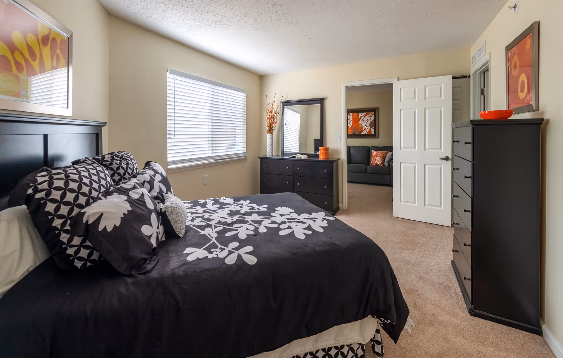 A bedroom with a black bed frame and black bedding featuring white floral patterns. The room has a window with blinds, a black dresser with a mirror, and another tall black dresser. There are two colorful abstract paintings on the walls. The door is open, showing a glimpse of a living room with a gray sofa and an orange pillow.