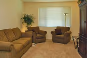 A cozy living room with a brown patterned sofa, two matching armchairs, a tall floor lamp, a potted plant in the corner, and a large window with closed blinds. The room has beige carpet and light-colored walls.