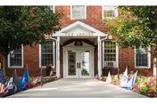 Brick building entrance with white porch roof, flags along the walkway, and trees framing the scene.