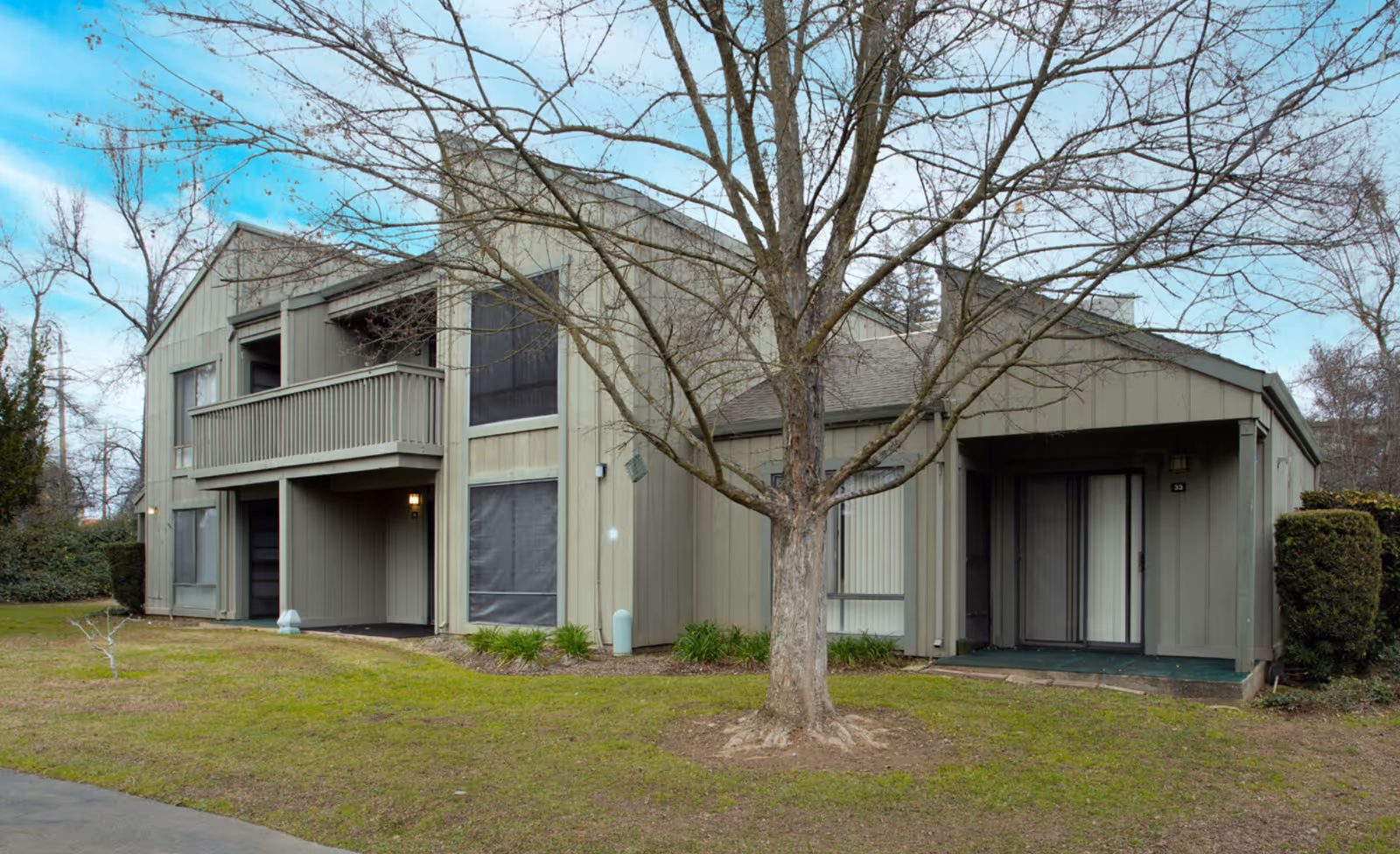Exterior view of a two-story residential building with a balcony, sliding glass doors, and a leafless tree in front on a grassy lawn under a partly cloudy sky.