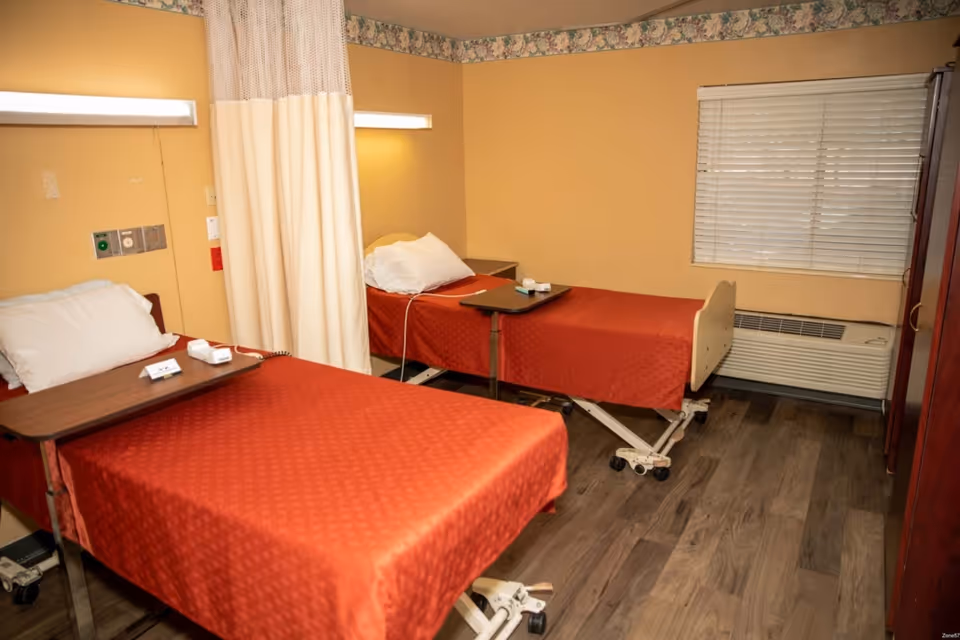 A room with two hospital-style beds covered with red bedspreads, each equipped with an overbed table. The room has beige walls, a window with closed blinds, a floral border near the ceiling, and wood-look flooring. A privacy curtain is partially drawn between the beds.