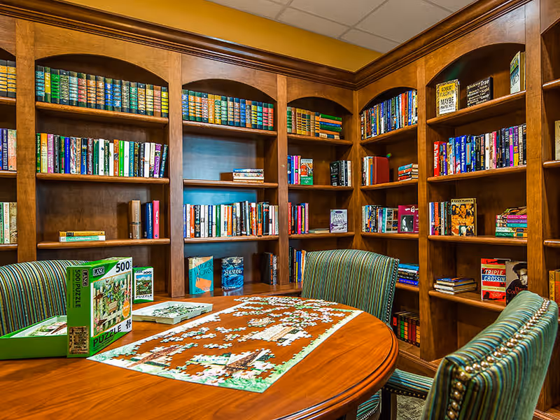 A cozy library-style room with wooden bookshelves, a round table holding a partially completed jigsaw puzzle and striped upholstered chairs.