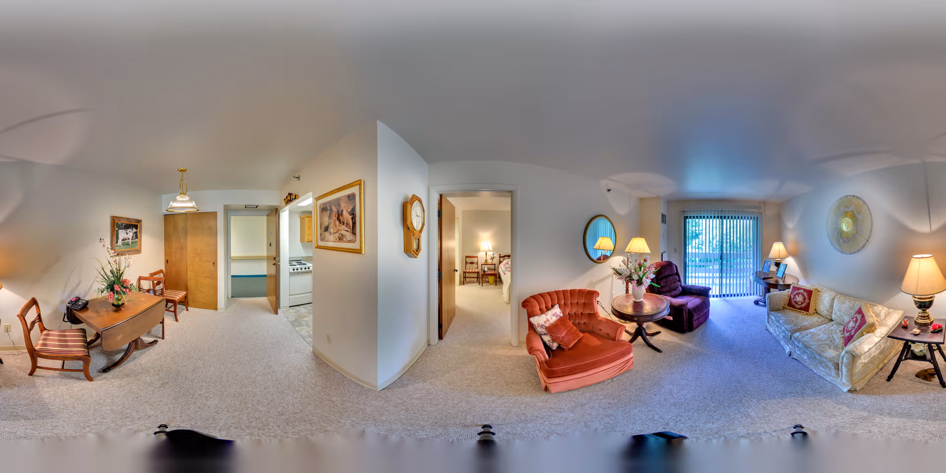 A panoramic view of a retirement community apartment interior showing a living room and dining area. The living room has a cream-colored sofa with red pillows, a red armchair, a purple recliner, a round wooden table with a flower vase, and several lamps providing warm lighting. Sliding glass doors with vertical blinds lead to an outdoor area. The dining area features a wooden table with two chairs, a hanging light fixture, and a framed picture on the wall. The kitchen is visible through an open doorway, and another room with a bed can be seen through a second doorway. The walls are painted white, and the floor is carpeted in a light color.