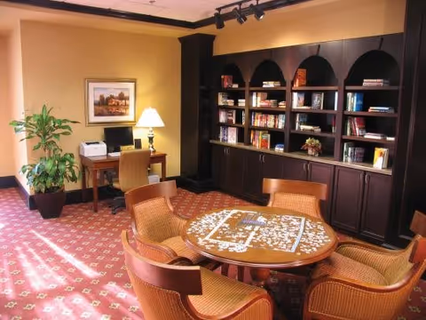 A cozy room with a round wooden table in the center surrounded by four wicker chairs. The table has a partially completed jigsaw puzzle on it. Behind the table is a large dark wooden bookshelf filled with books and decorative items. To the left, there is a small desk with a computer, printer, and a lamp, next to a potted plant. The room has warm yellow walls and a patterned red carpet.