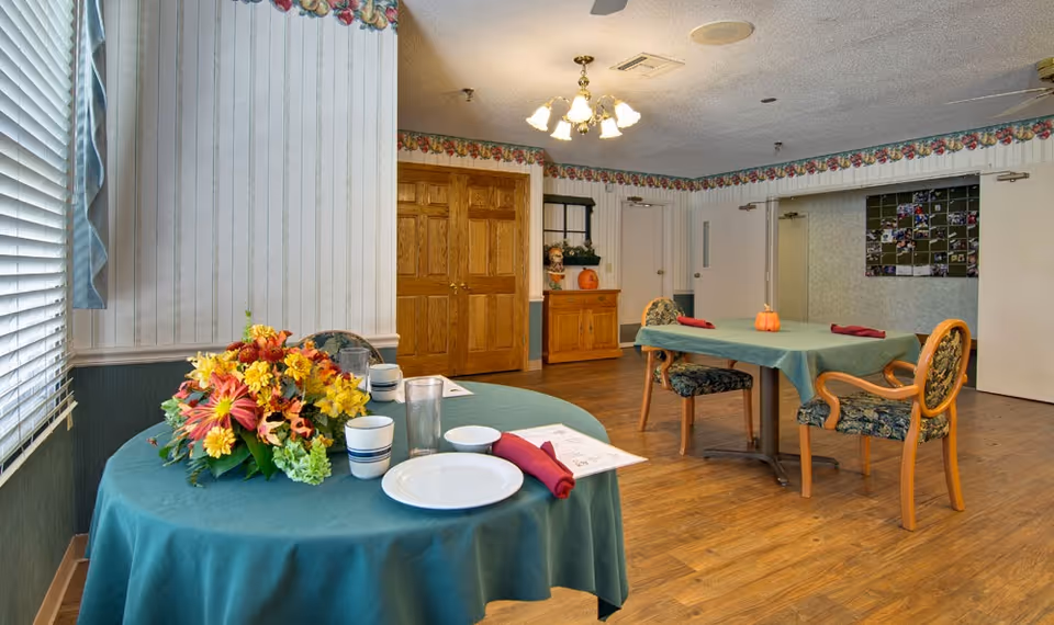 A dining room with two tables covered in teal tablecloths, each set with plates, cups, napkins, and menus. One table has a colorful floral centerpiece, and the other has small pumpkin decorations. The room has wooden floors, floral wallpaper border, wooden double doors, and a bulletin board on the far wall.
