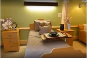 A neatly made single bed with gray bedding and multiple pillows in a room with light green walls. There is a wooden nightstand on each side of the bed, one with a decorative clock and flower artwork, and the other with a vase and decorative items. A tray with a bowl and plate is placed on the bed. A mustard-colored armchair is visible in the background.