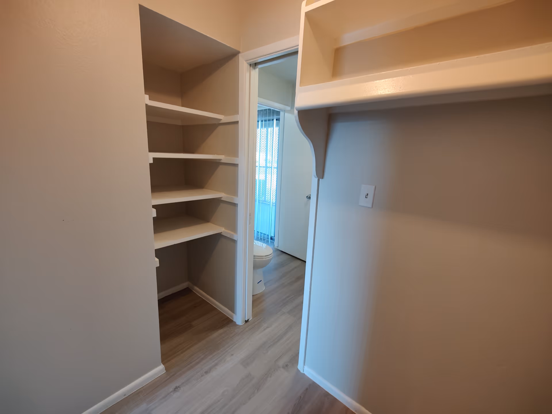 Interior view of a small room with built-in white shelves on the left and right walls. The floor is light wood laminate. Through an open doorway, a bathroom is visible with a toilet and vertical blinds covering a window or door.