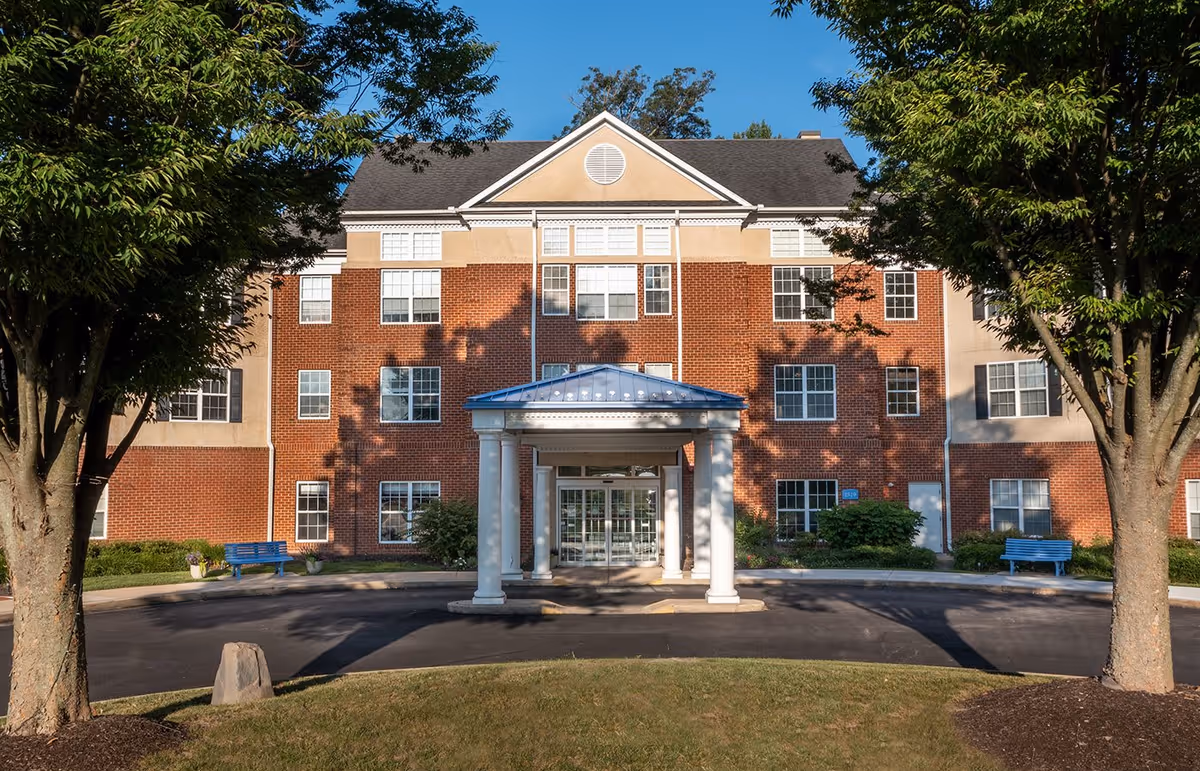 Front exterior view of a three-story brick building with a covered entrance supported by white columns. The building is surrounded by trees and has a paved driveway with blue benches on either side near the entrance.