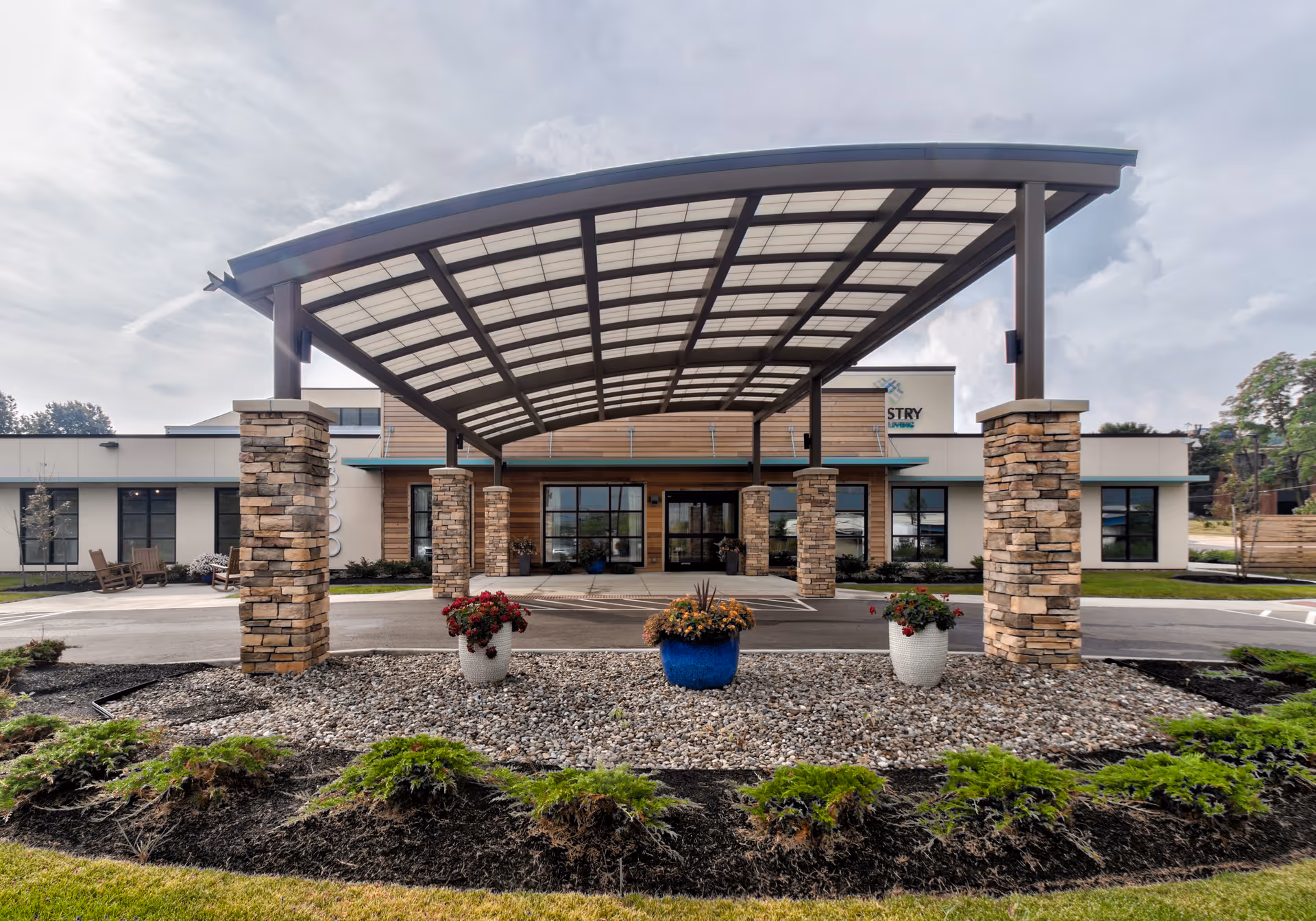 Front exterior view of a senior living facility with a large covered entrance supported by stone pillars. There are three flower pots with colorful flowers placed on a bed of rocks in front of the entrance. The building has large windows and a modern design with wood and stone accents.