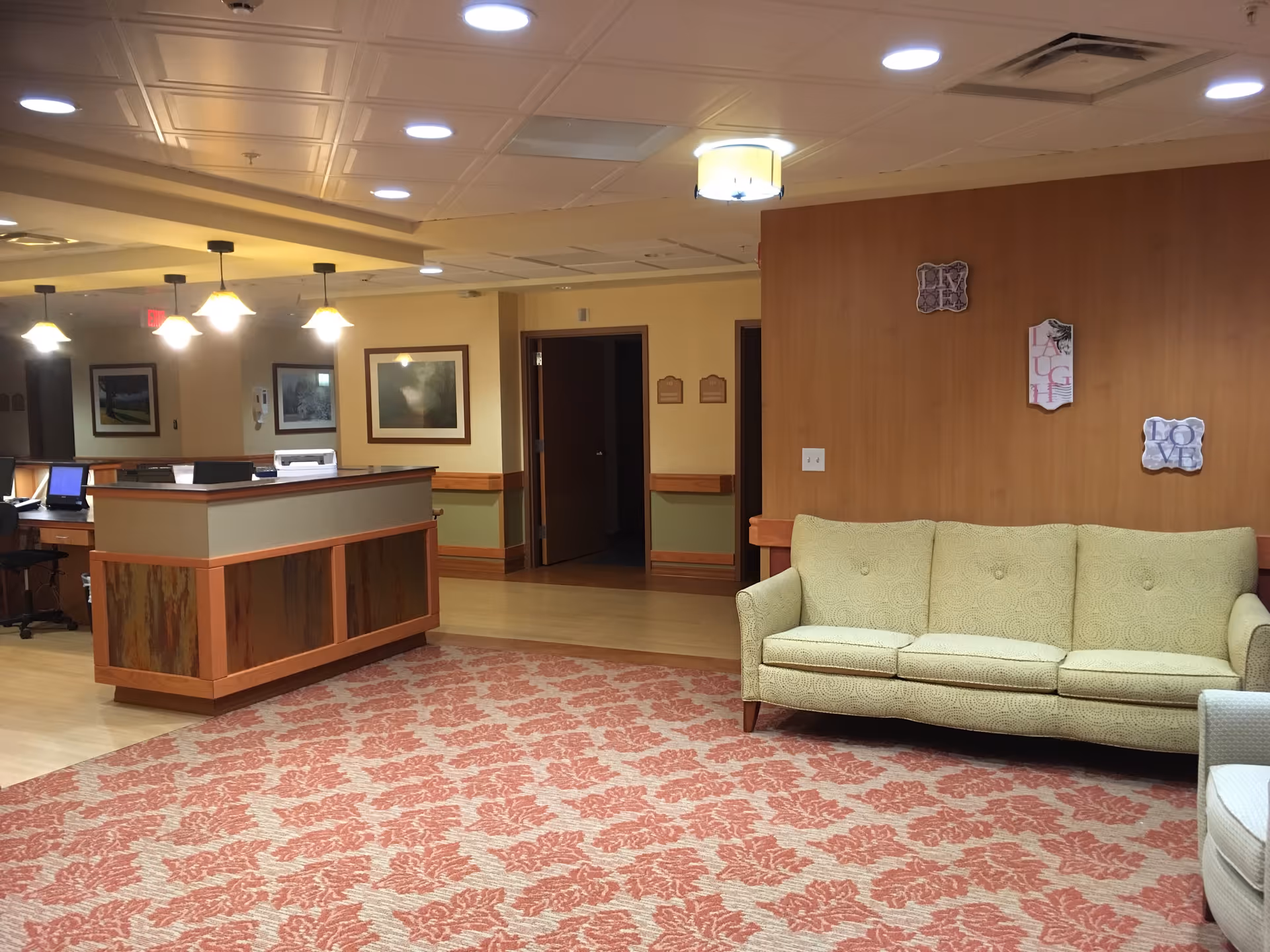 Interior view of a senior living facility lobby area with a wooden reception desk, a green patterned sofa, a red and beige floral carpet, framed pictures on the walls, and warm lighting from ceiling fixtures.